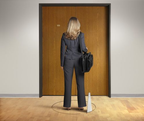 A rear view of a businesswoman standing with a shoulder bag waits in front of a closed office door while someone below her is attempting to cut out the floor from under her using a saw to cut through the hardwood floor.